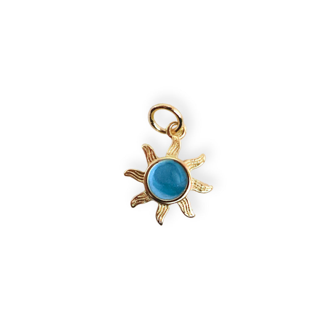 Dije en forma de sol de chapa de oro con centro esmaltado en azul sobre fondo blanco