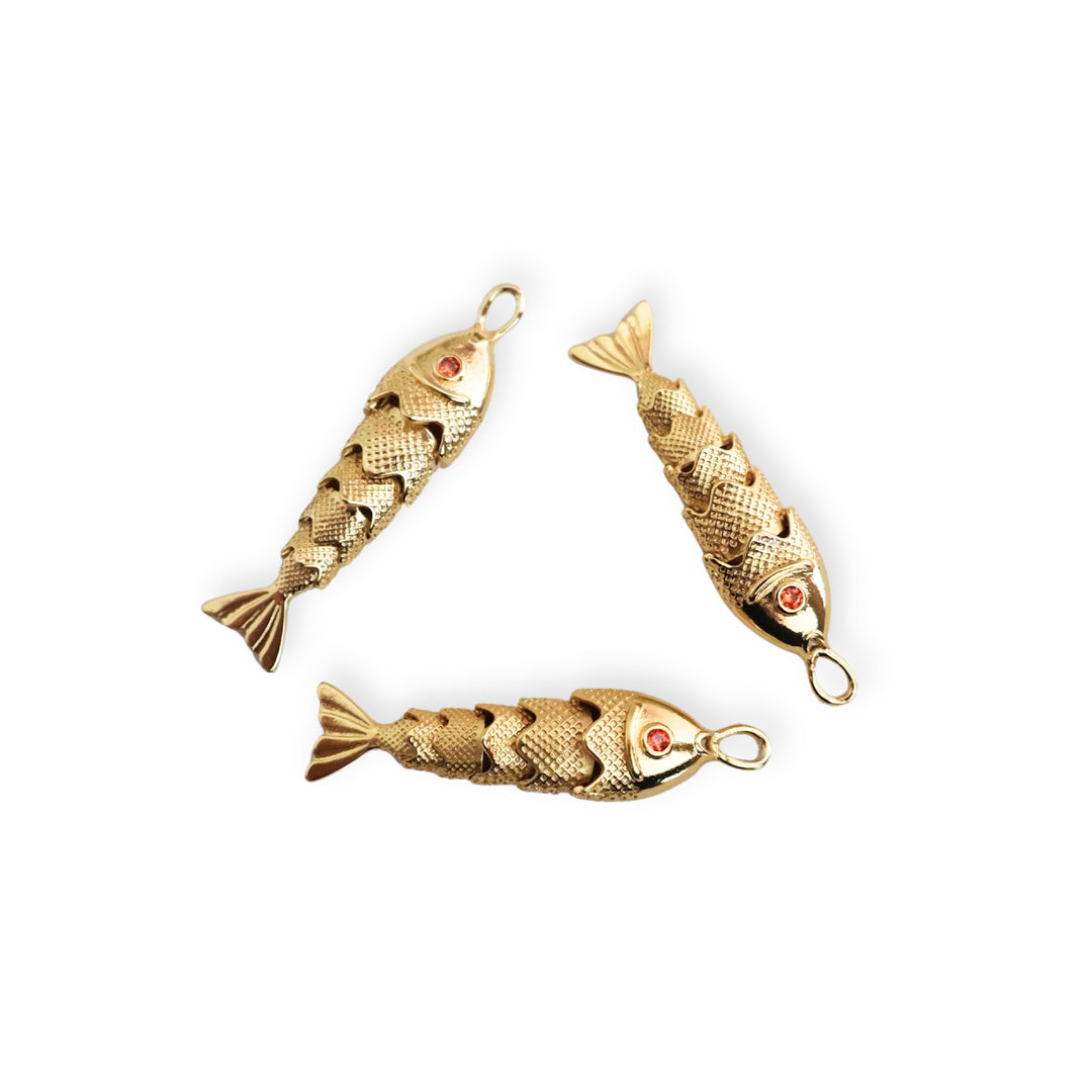 Detalle de dijes dorados en forma de pescado con zirconia roja en el ojo y cuerpo articulado sobre fondo blanco