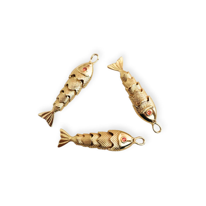 Detalle de dijes dorados en forma de pescado con zirconia roja en el ojo y cuerpo articulado sobre fondo blanco
