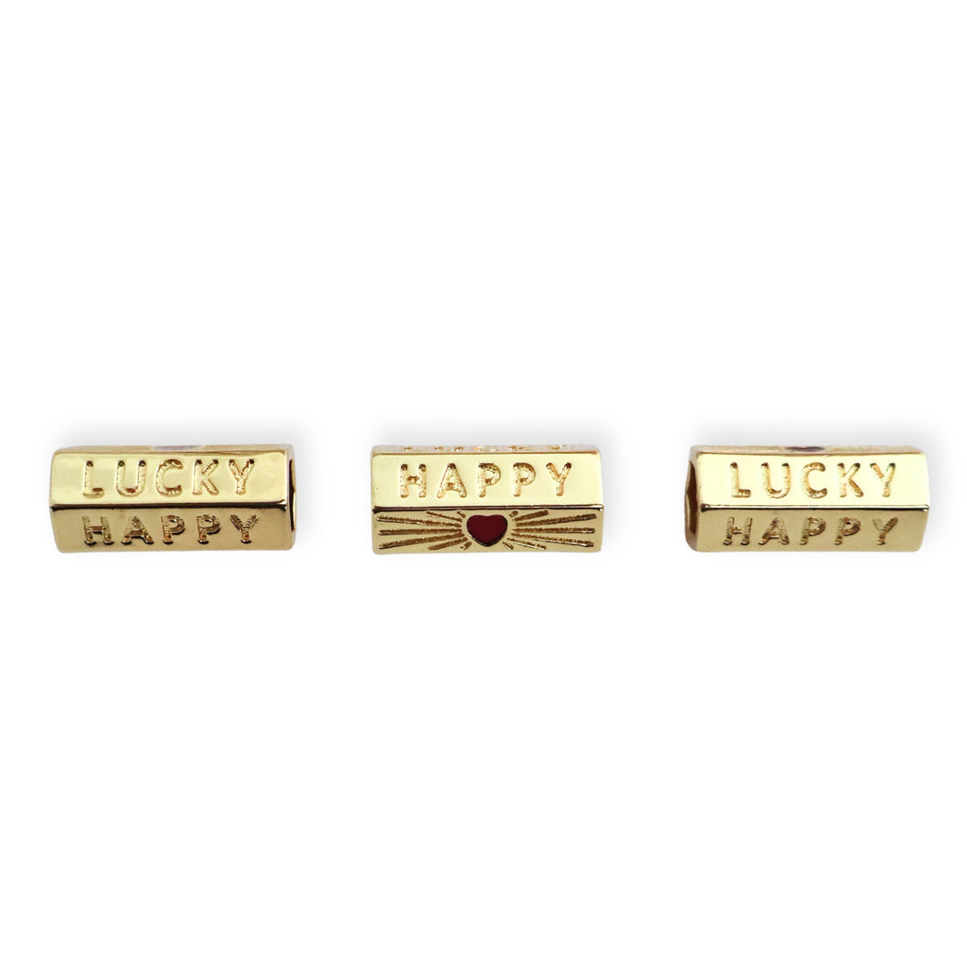Toma a detalle de separadores de chapa de oro con grabado "Happy", "Lucky" y corazón rojo esmaltado sobre fondo blanco