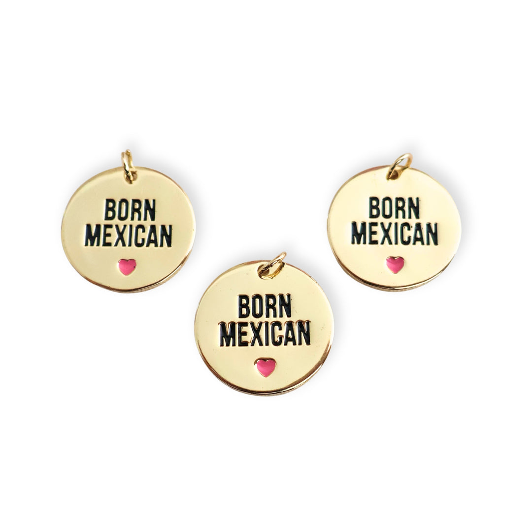 Detalle de dijes redondos de chapa de oro con frase "BORN MEXICAN" y corazón rosa esmaltado sobre fondo blanco
