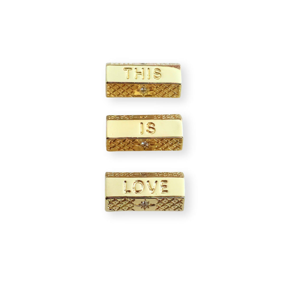 Toma a detalle de separadores de chapa de oro con grabado "THIS IS LOVE" y zirconia sobre fondo blanco