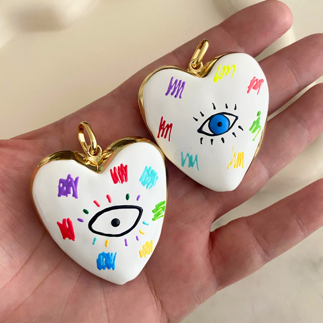 Dije grande de corazón esmaltado blanco con ojo central y detalles multicolor en chapa de oro en mano.