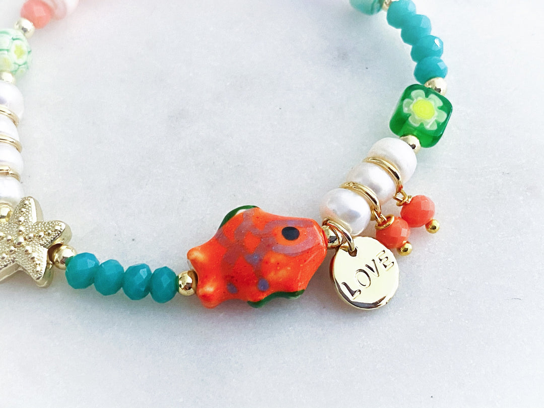 Detalle de los dijes de estrella dorada y pez de cerámica en la pulsera Bruma con cristales y perlas.