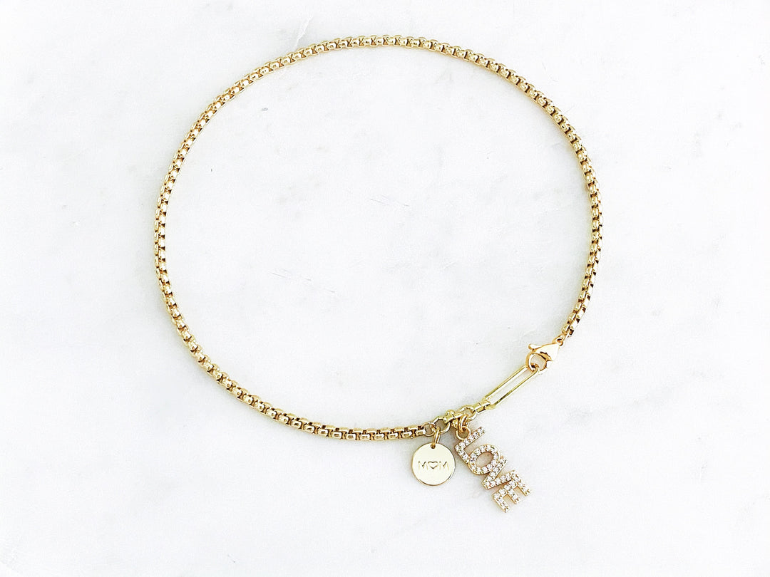 Detalle del dije dorado “mamá” con zirconia, placa “love” y corazón de concha en el collar Orígen.