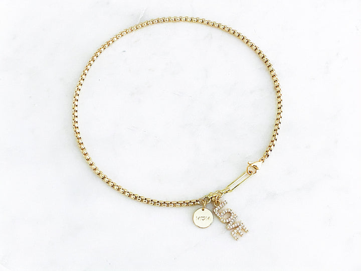 Detalle del dije dorado “mamá” con zirconia, placa “love” y corazón de concha en el collar Orígen.