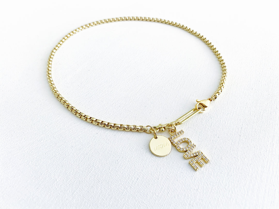 Detalle del dije dorado “mamá” con zirconia, placa “love” y corazón de concha en el collar Orígen.
