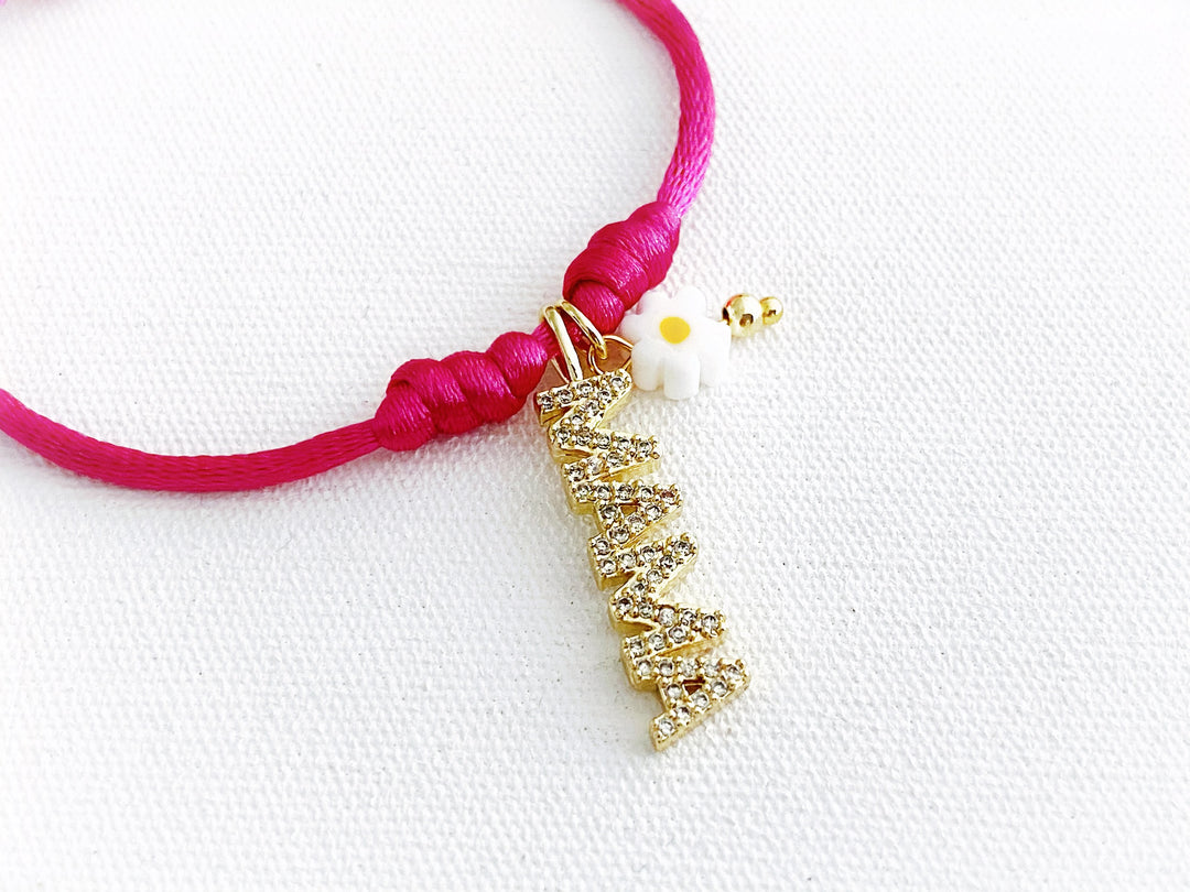 Detalle de pulsera ajustable de hilo fucsia con dije dorado “mamá” y flor metálica de murano, vista completa.